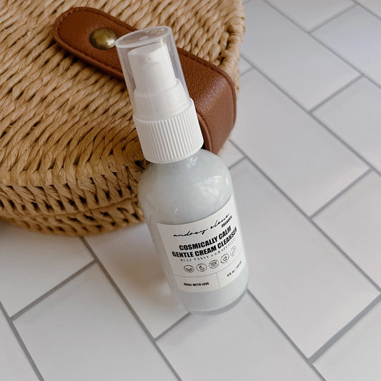 A glass pump bottle of Blue Tansy + Grapeseed Gentle Cream Cleanser placed on a white tiled surface next to a wicker bag.