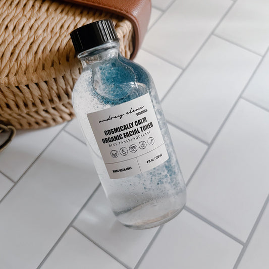 A clear glass bottle of organic facial toner with blue contents visible through the bottle, labeled 'Audrey Elene Organic Blue Tansy x Squalane', placed on a tiled surface next to a woven basket.
