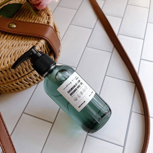 A glass bottle with a pump dispenser containing organic body oil, placed on a tiled surface next to a wicker bag and a brown belt.