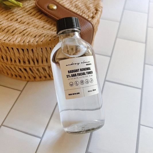 A clear glass bottle labeled 'Glycolic Acid x Lactic Acid 5% AHA Facial Toner' from Audrey Elene Organics, placed on a tiled surface next to a wicker object, presumably a handbag.