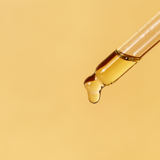 A glass dropper with a few drops of facial oil hanging from it, against a light background.